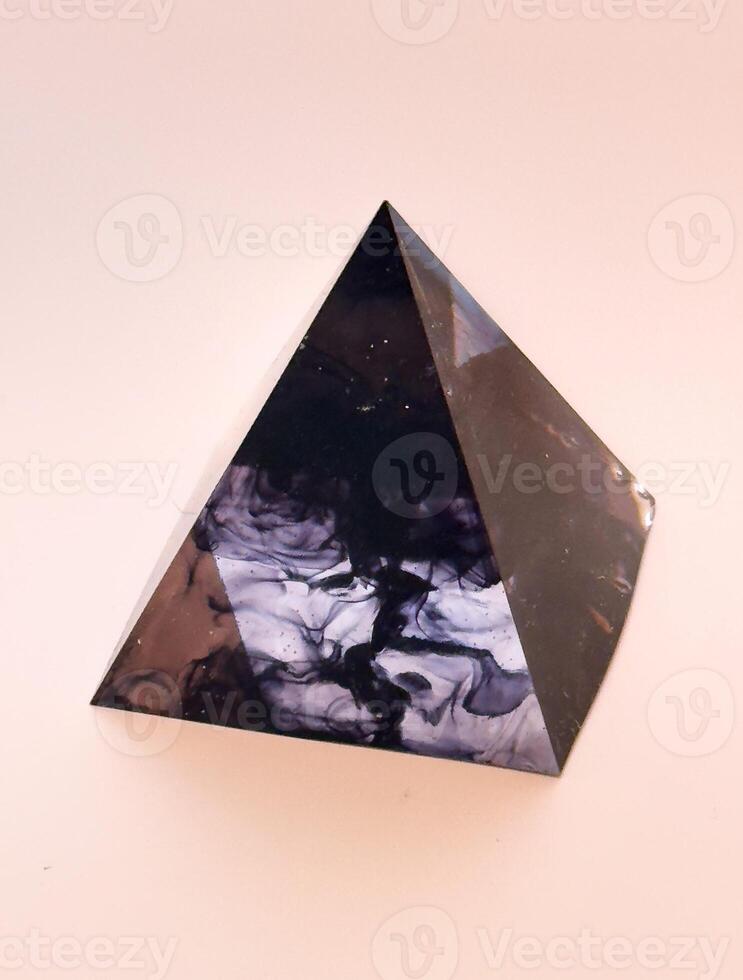 An intriguing eye-level shot captures a polished, dark pyramid with captivating internal swirls and patterns, hinting at a natural stone or resin. The object rests on a soft, light-colored background photo