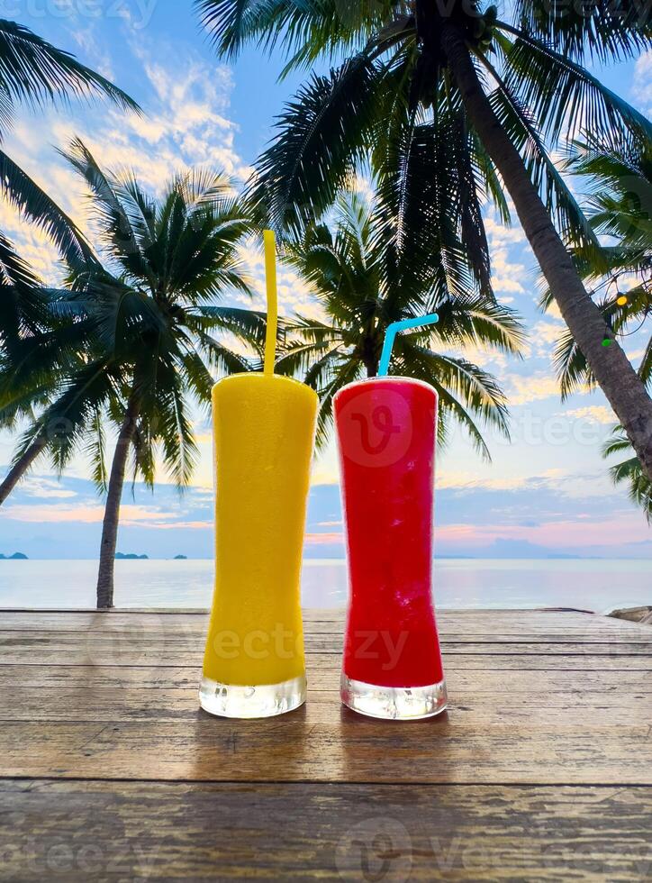 Two vibrant, tall glasses filled with colorful tropical drinks, one bright yellow and one vivid red, stand on a rustic wooden table. In the background, silhouetted palm trees frame a serene beach photo