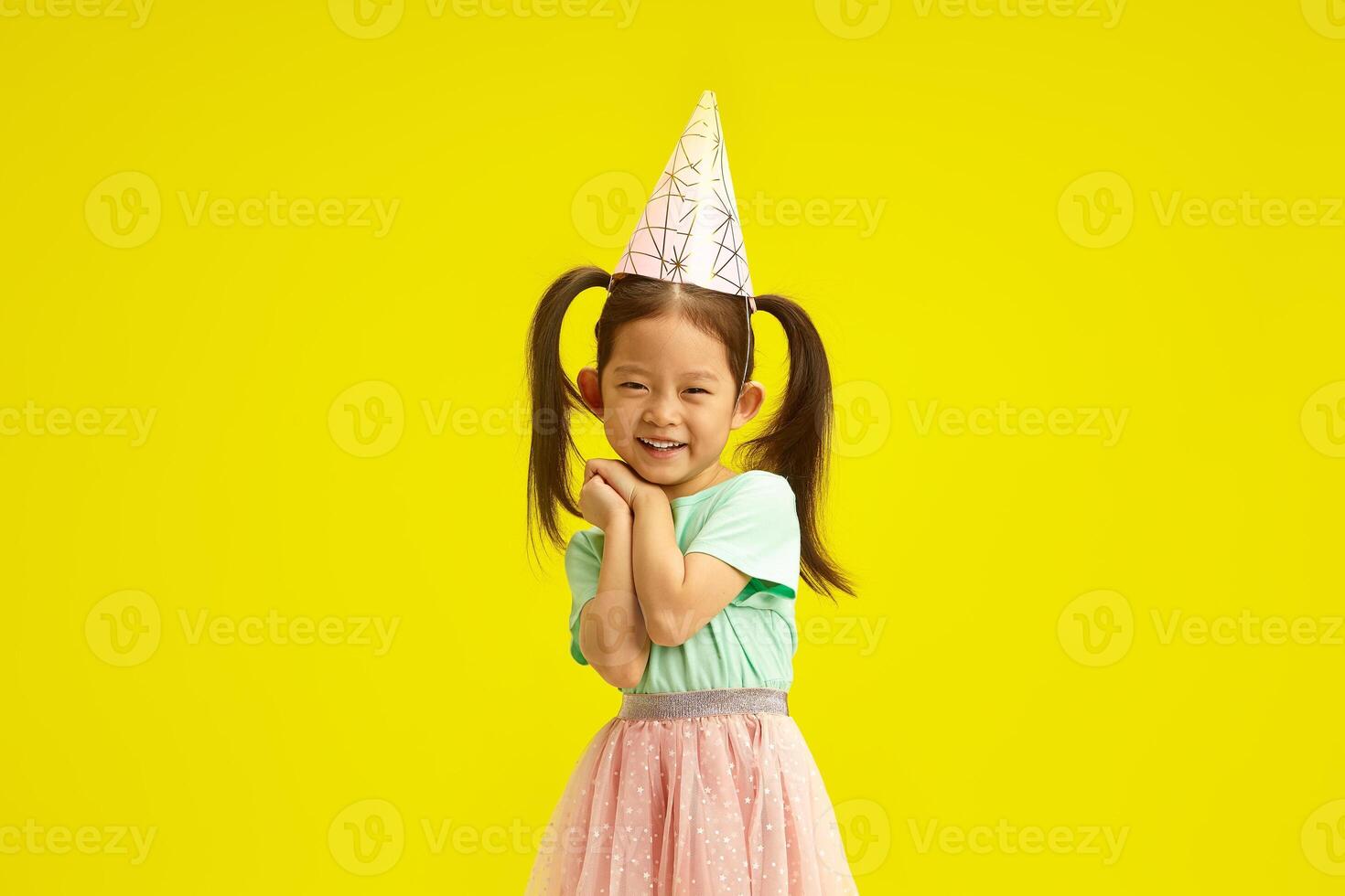 Asian little girl in party hat smiling coyly, cute placed palms of hands in front of herself face, looks like as if begging for a desired gift or makes a wish on herself birthday, stands over yellow photo