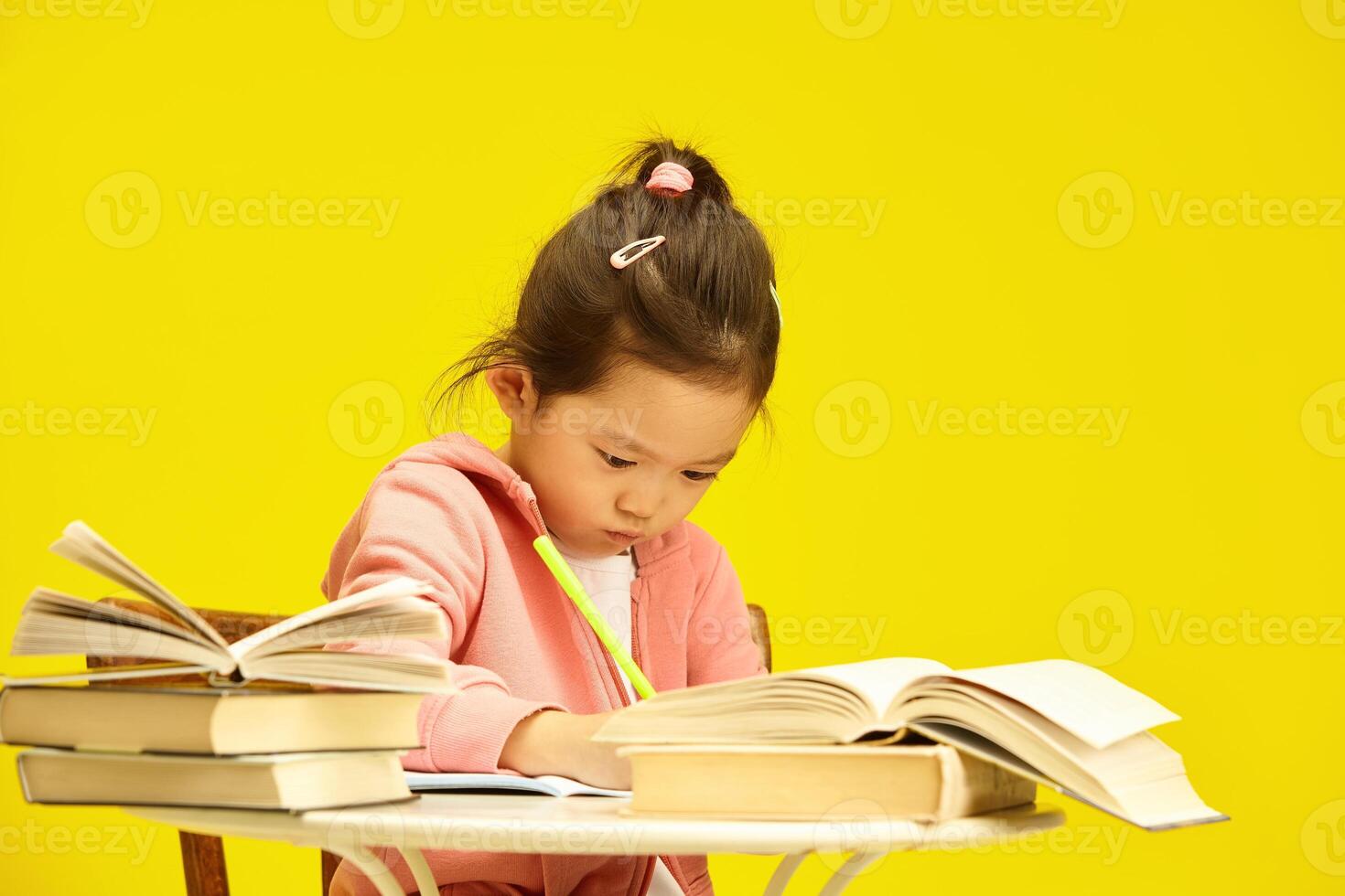 Studious Asian preschooler child girl deep in study amidst a pile of books sitting at table over sunny yellow isolated background, writing somethnig in copybook photo