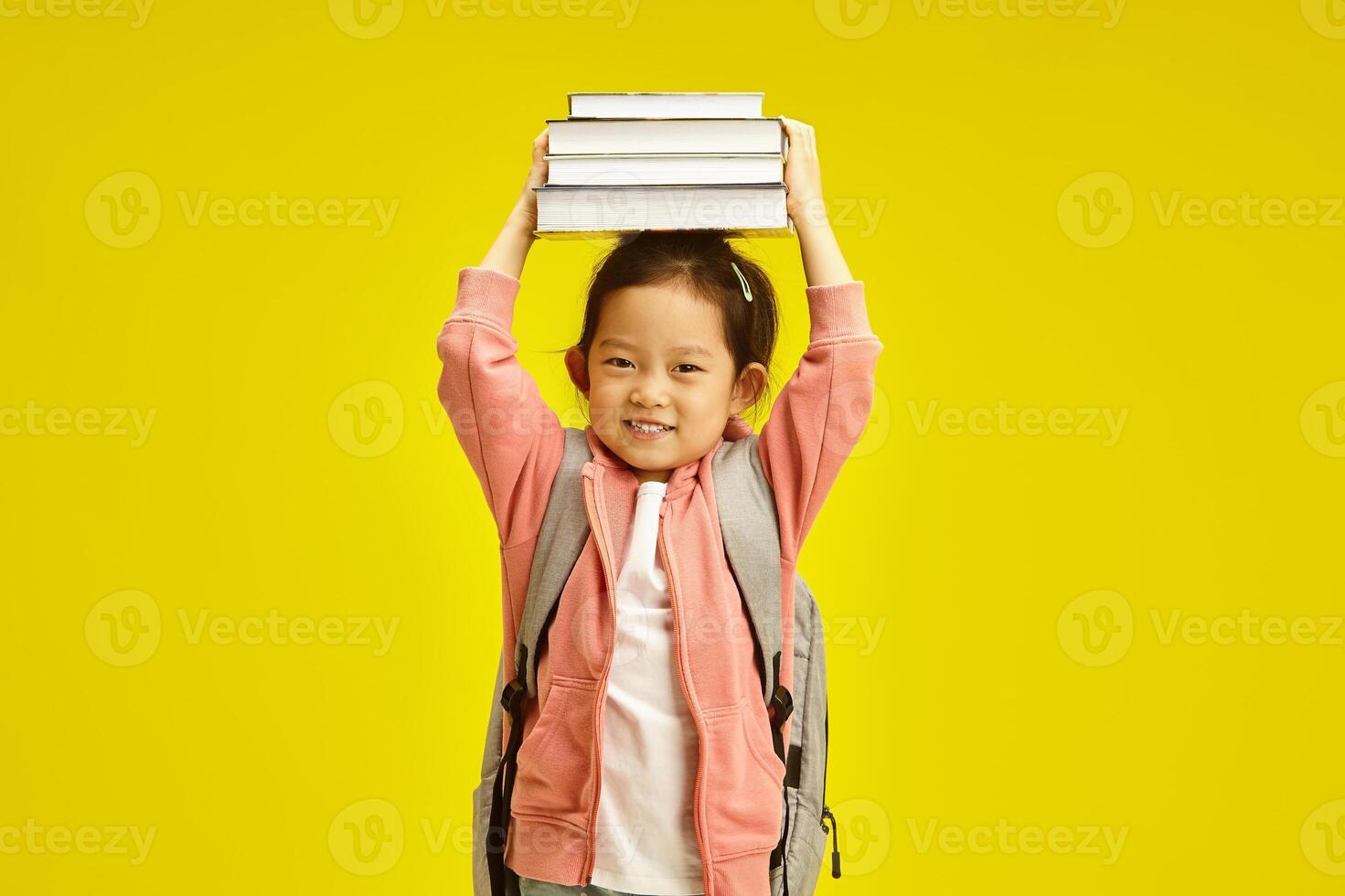 Sweet first grader asian ethnicity schoolgirl holding books above head, cheerful smiling standing with backpack on her shoulders over yellow isolated background. photo