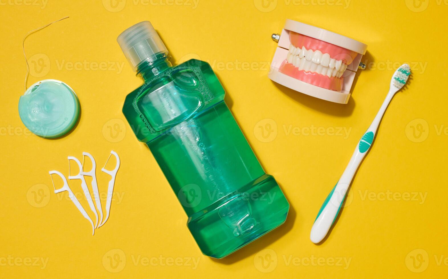 Dental floss in a case, green mouthwash bottle, jaw model, toothbrush and plastic flossers arranged on a bright yellow background. photo