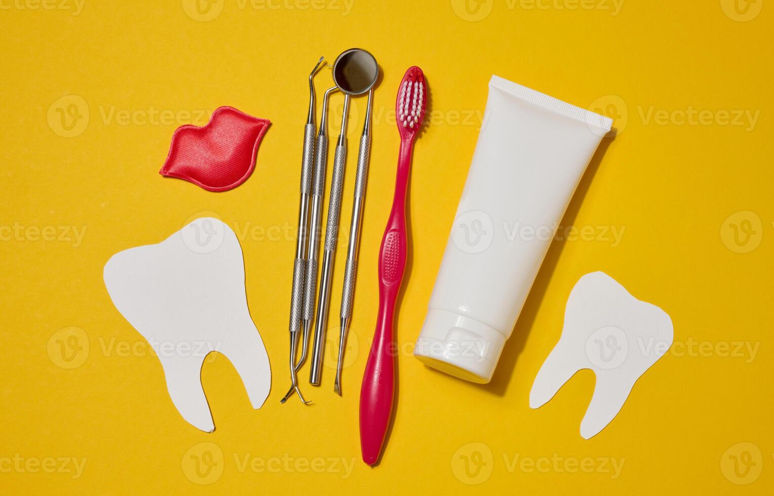 Red toothbrush, white tube of toothpaste, red decorative lips, dental instruments, dental floss in a case and white paper teeth cut out on a yellow background. photo