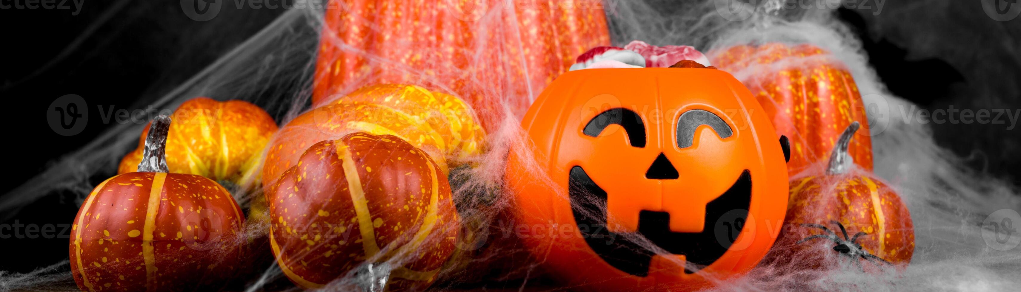 Dark halloween banner with trick or treat concept. Jack-o-lantern bowl full of sweets near a pile of pumpkins covered in spiderweb photo