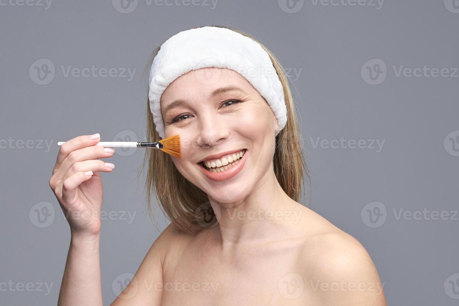 Smiling caucasian female adult applying makeup with brush on cheek with headband on gray background. photo