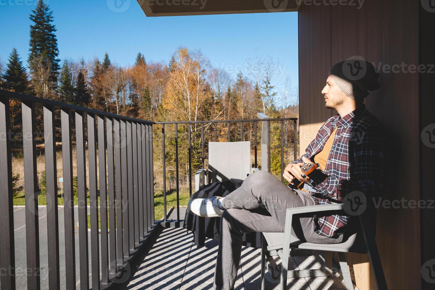 Musician sits on a sunny balcony, immersed in playing the ukulele. Surrounded by autumn trees, he enjoys the peaceful moment outdoors, lost in his thoughts and music photo