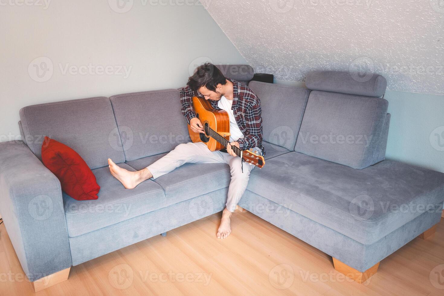 Man sits comfortably on a gray couch. Relaxed young man plays his guitar with passion photo