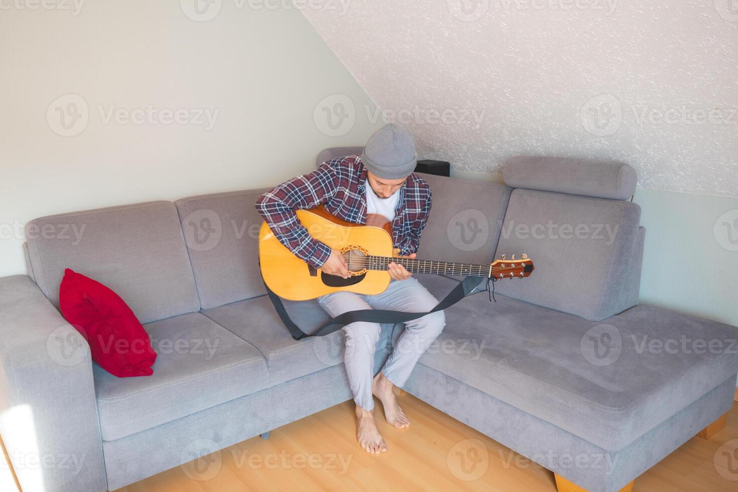 Barefoot musician sits comfortably on a gray couch. Close-up of guitarist in mid-performance at home photo