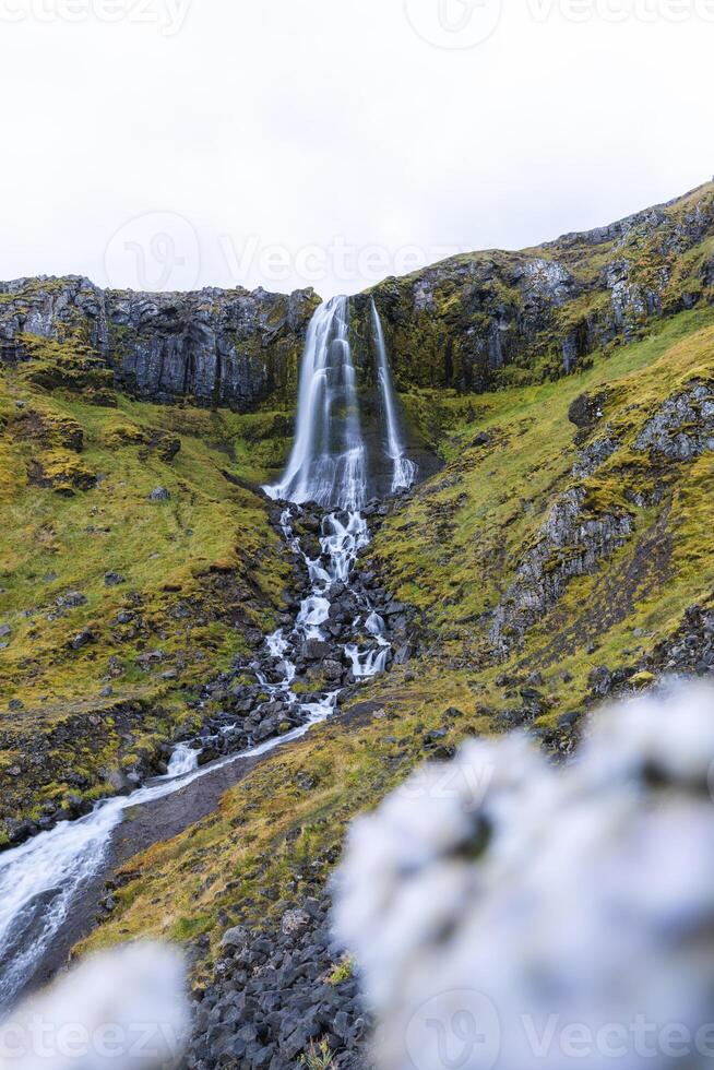 Baejarfoss waterfall elegantly down mossy cliffs in Olafsvik, Iceland, surrounded by lush green slopes and volcanic rock in a serene coastal landscape photo