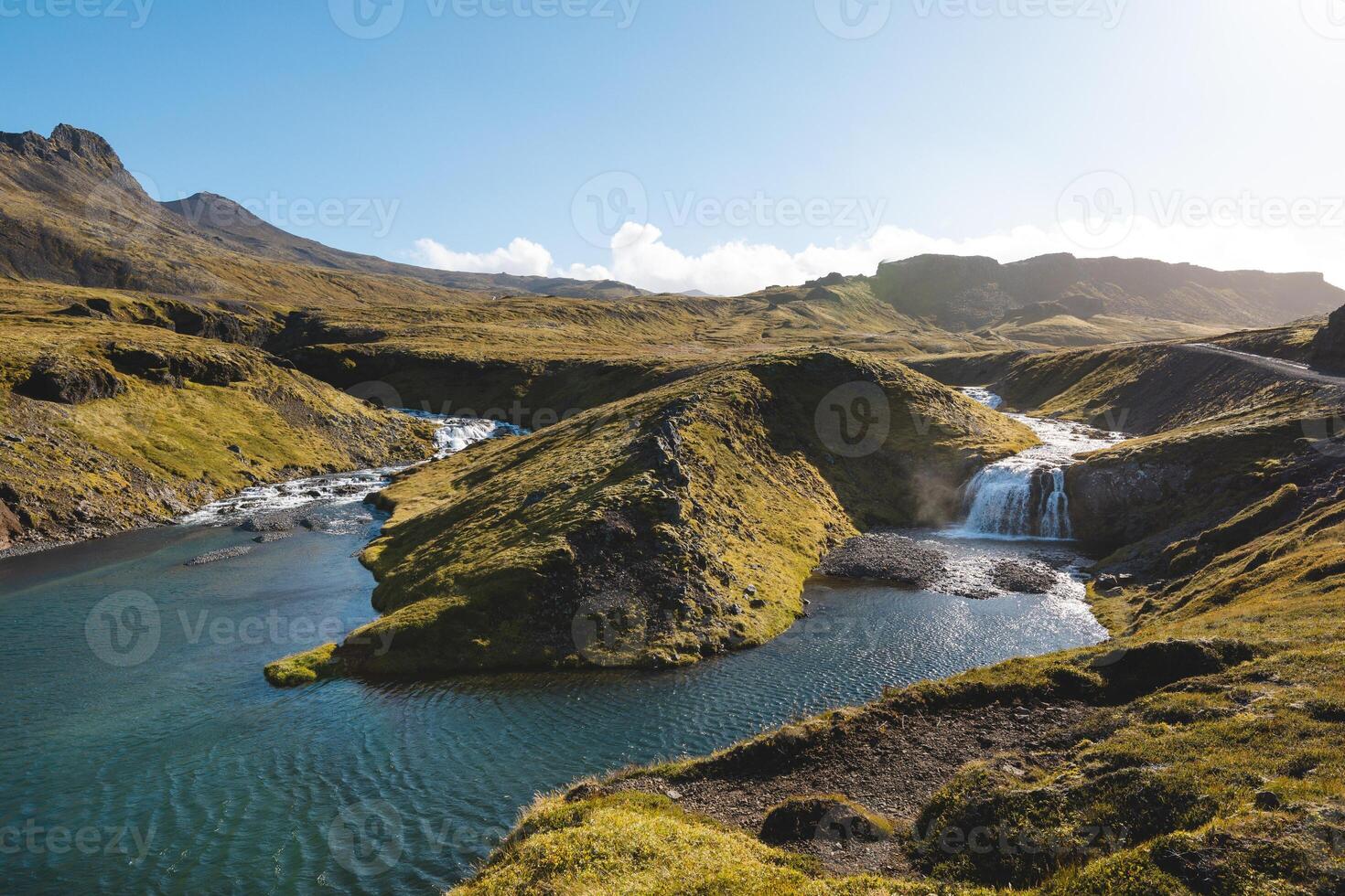 Serene Icelandic stream winds through mossy hills, forming small waterfalls and crystal-blue pools under clear skies, surrounded by untouched volcanic landscapes photo