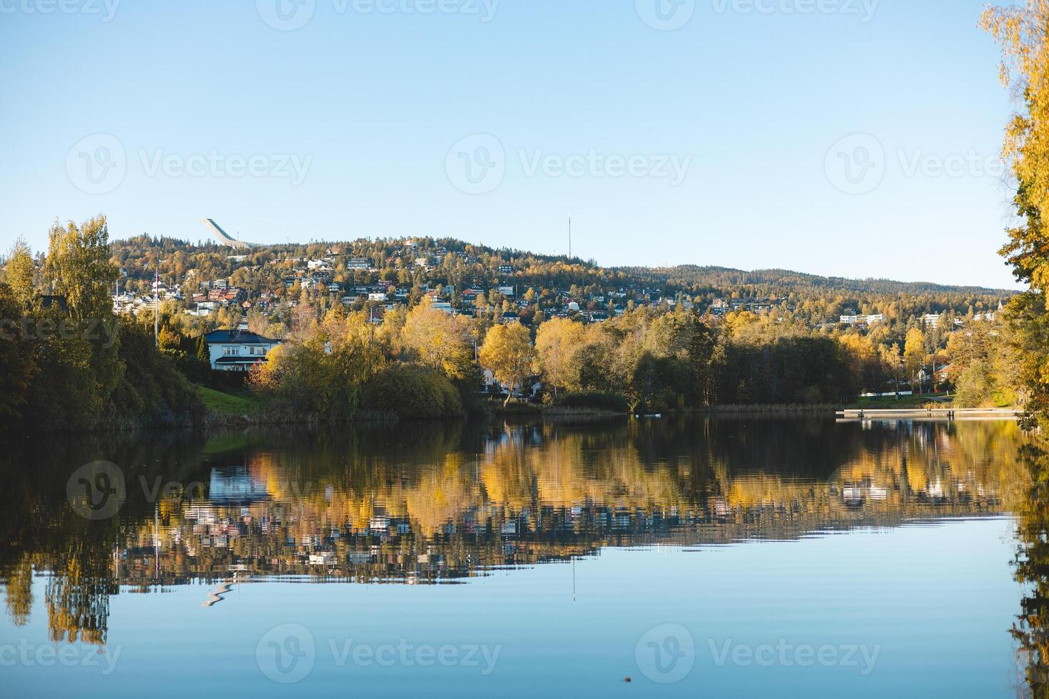 Autumn reflections dance on the still waters of Holmendammen Lake in Oslo, surrounded by colorful trees and hillside homes under a crisp, cloudless sky photo