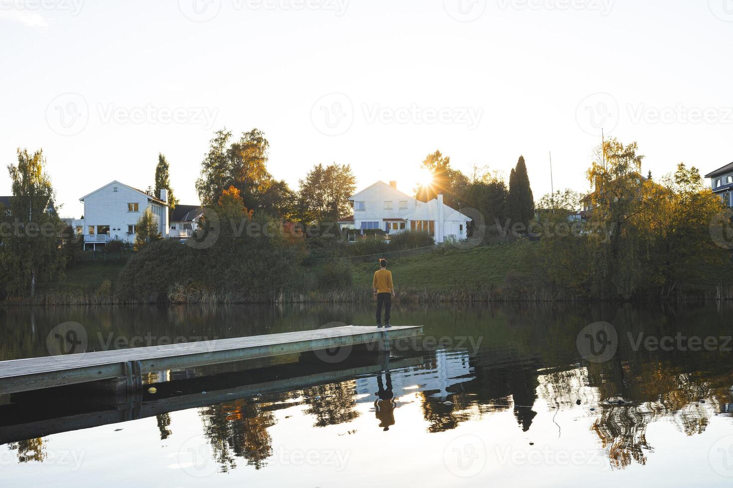 Boy walks on pier an Oslo lake, Holmendammen as the sun sets behind charming houses, capturing a peaceful moment of reflection and stillness in Norway autumn magic photo