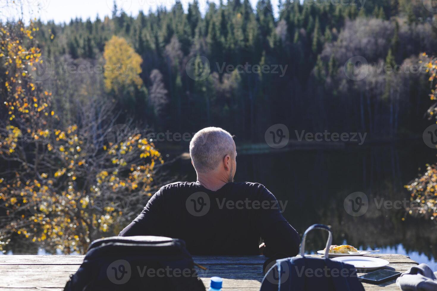50 years old man relaxes by a forest lake in Oslo Marka, enjoying lunch under golden trees and sunlight, surrounded by still water and deep Scandinavian calm photo