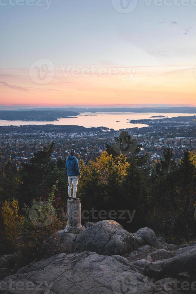 High above Oslo, a lone soul balances on stone, surrounded by golden trees and fading daylight, where forest stillness meets the endless horizon of the fjord photo
