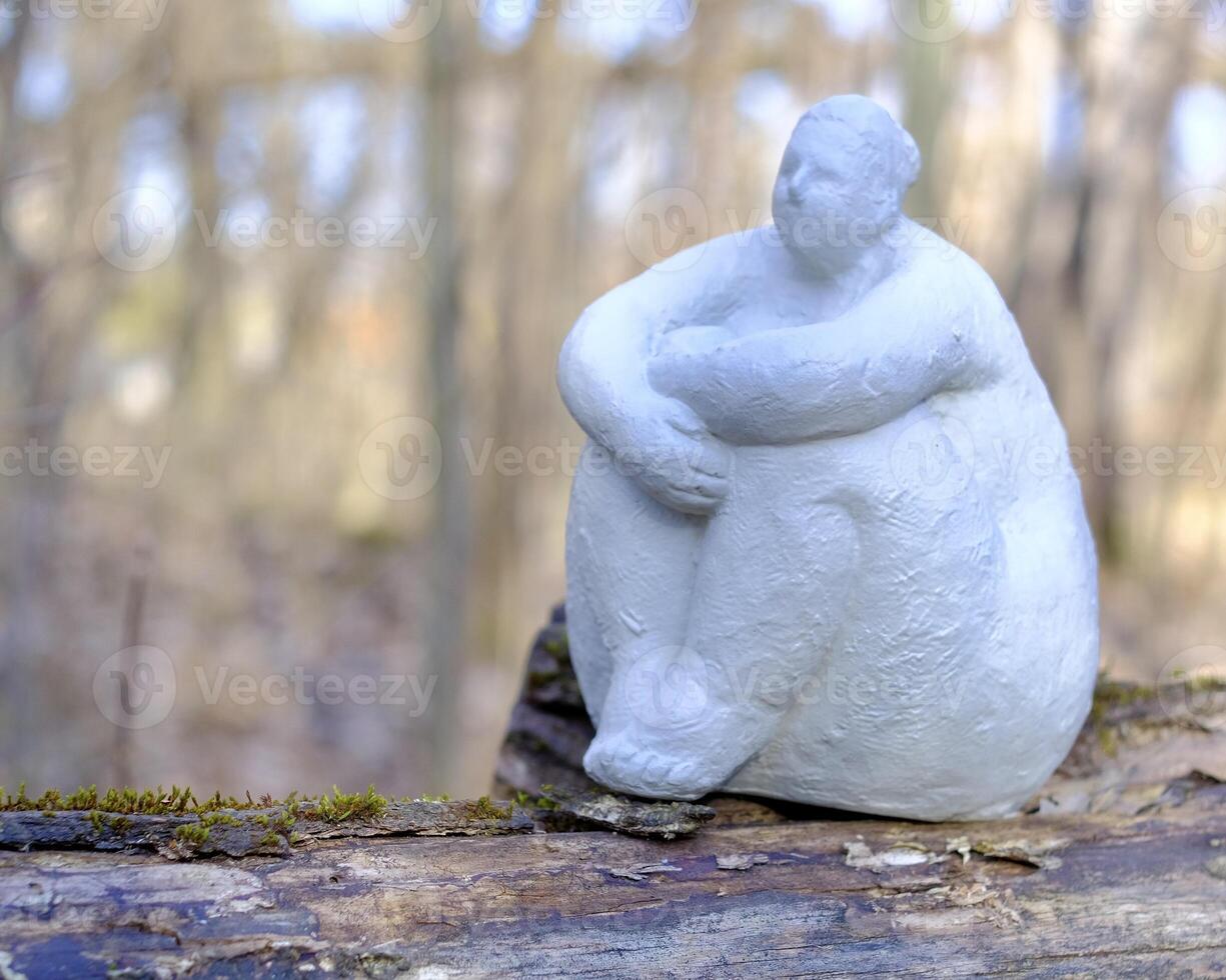 A plaster sculpture of a curvy, dreamy woman sitting on the trunk of a fallen tree, surrounded by the soft whispers of a blooming spring park photo