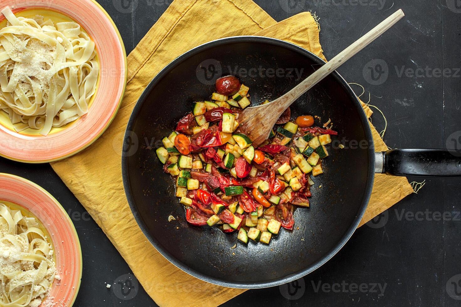 Overhead view of pasta dishes and wok with vegetables on a yellow cloth and a dark background photo