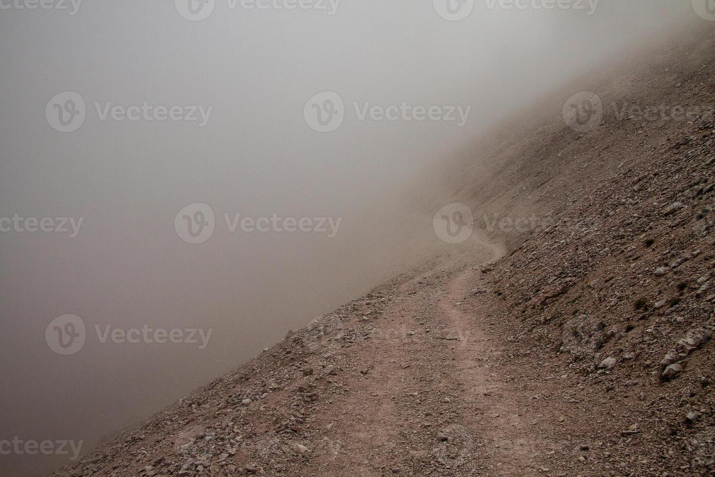 A view of a dirt path along a rocky slope covered in a thick layer of dense fog and mist air photo