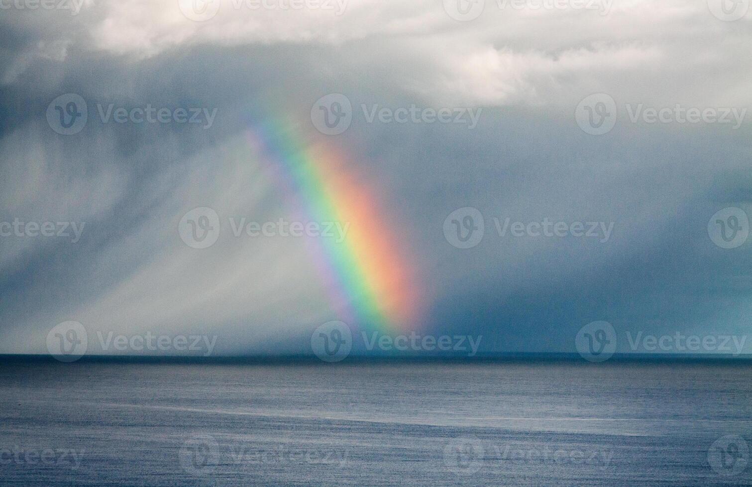 A rainbow over the ocean under a cloudy sky with visible rain and light breaking through the clouds photo