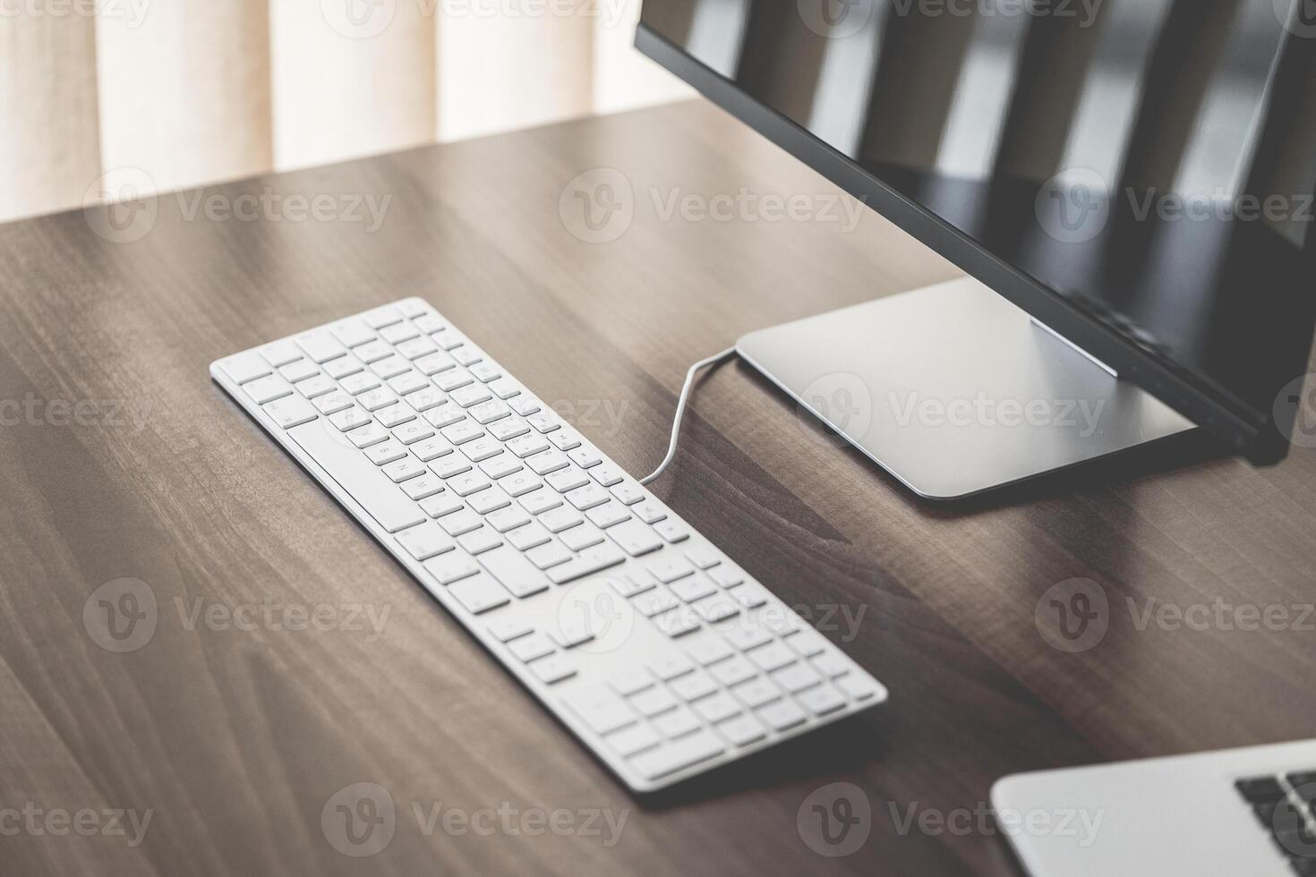A minimalist workspace featuring a keyboard, monitor, and laptop on a wooden desk surface near a window photo