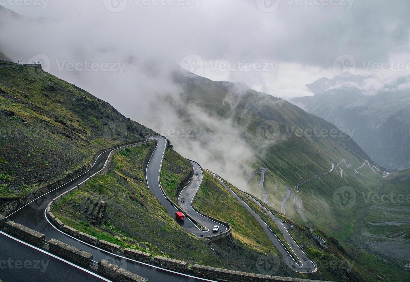 Winding mountain road disappearing into the clouds with cars on the road and green hillsides photo
