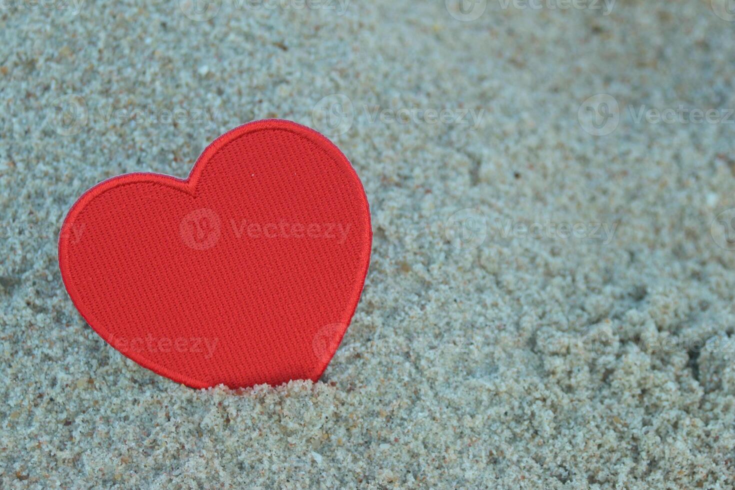 Close-up of red heart embroidery crafted from thread and fabric on fluffy beach sand. Heart placed on the left, creating space on the right for message or design. Valentine, romance vibe. photo