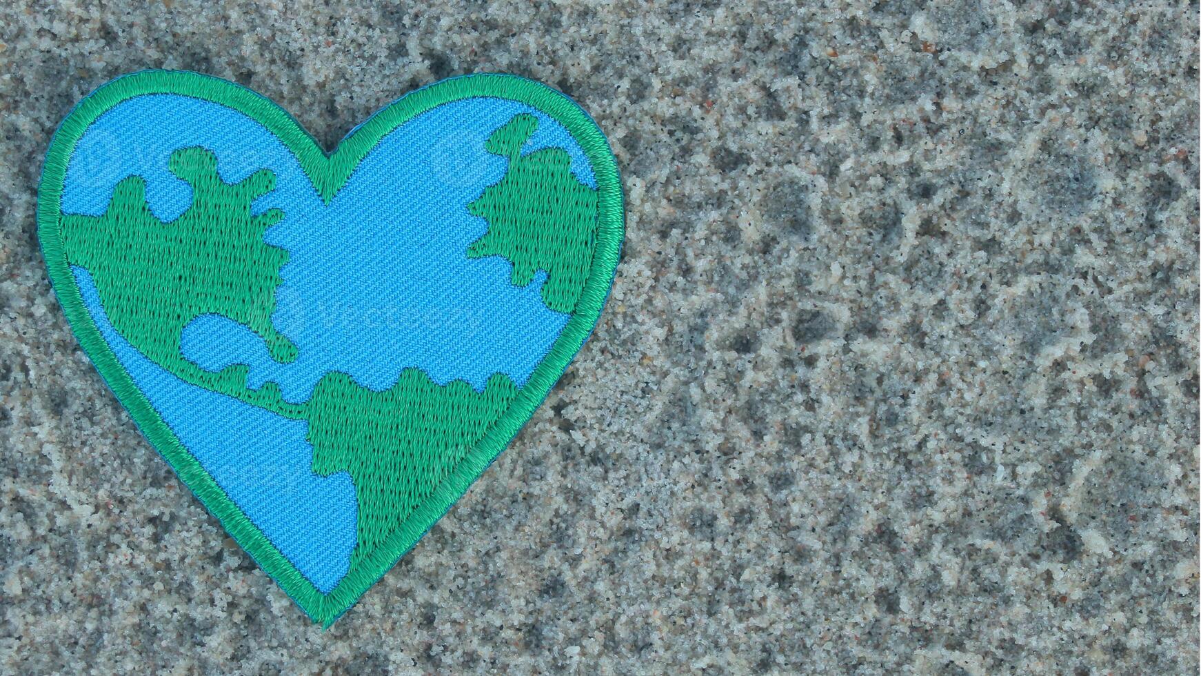 Green and blue Earth-patterned heart embroidery on sandy beach, positioned to the left with copy space on the right. Natural light, perfect for Earth Day and eco-friendly concepts. photo