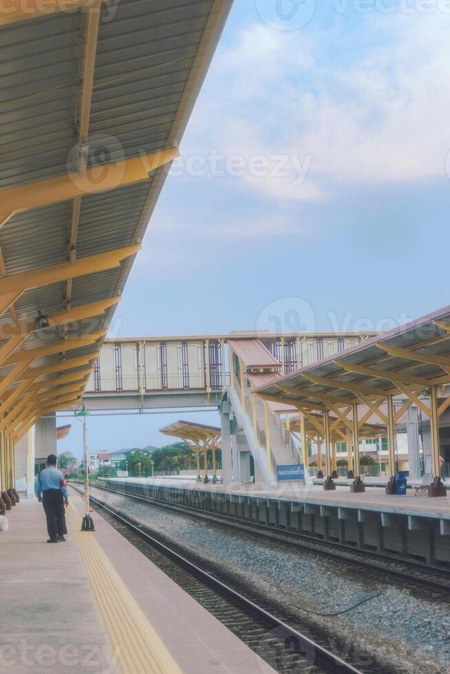 Ratchaburi Railway Station in Thailand, featuring dual train tracks, golden-yellow platform, and striking overhead crossing. Clear blue sky with soft clouds sets a perfect travel mood. photo