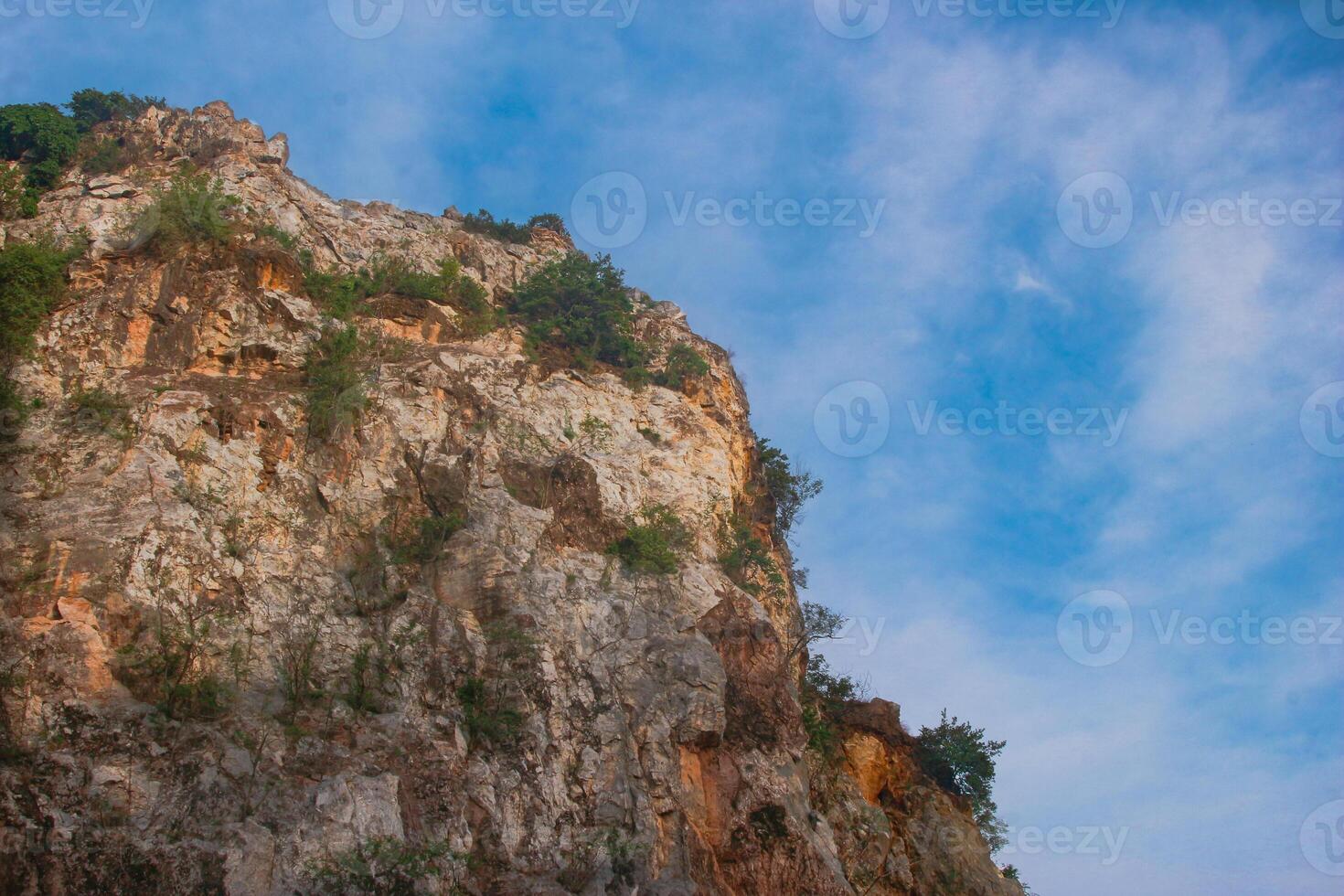Zoomed view of rocky mountain top with thin clouds hovering over the peak. Natural sunlight casts dramatic shadows on the rocky surface, framed by greenery. Southeast Asia atmosphere. photo