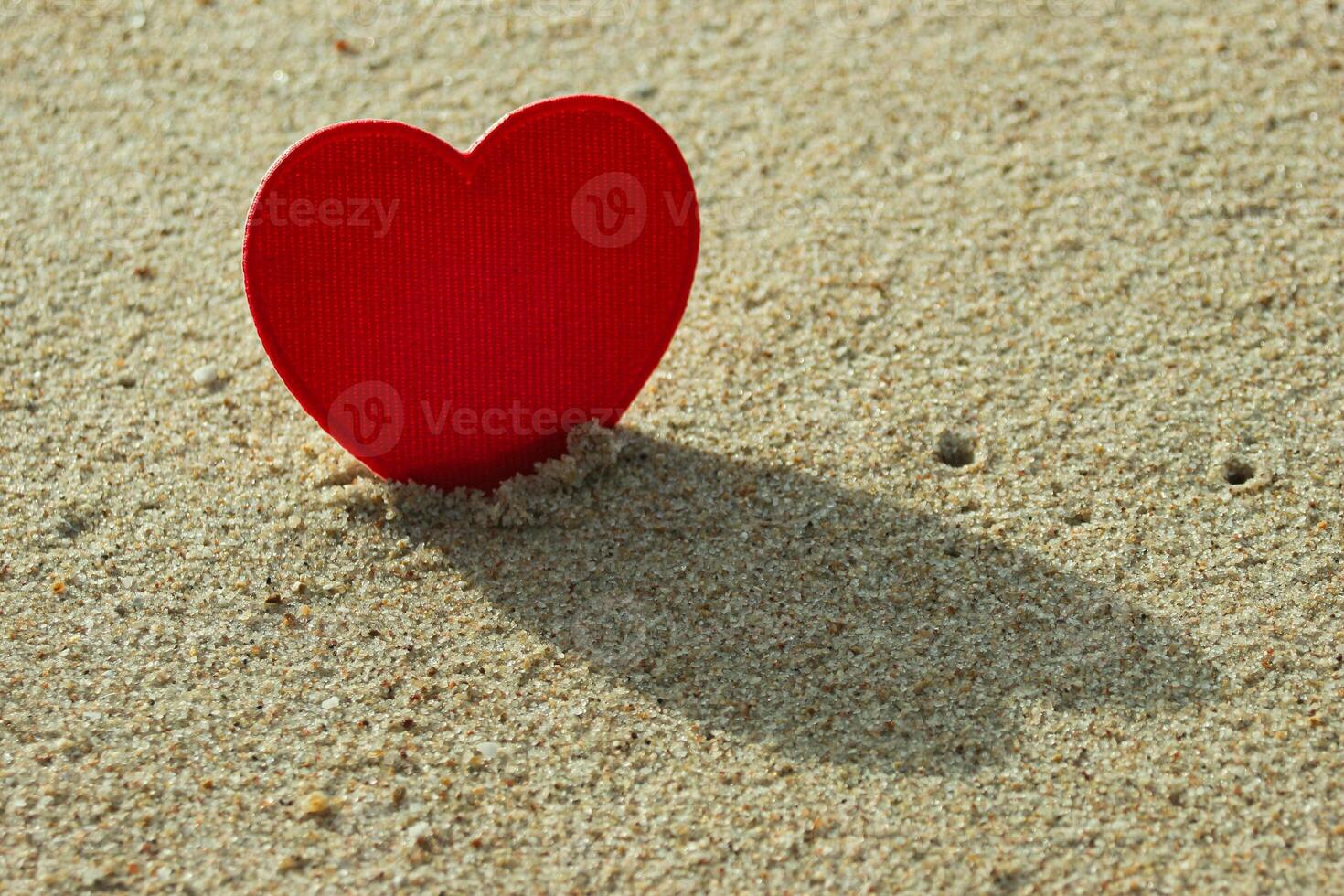 Close-up of red heart embroidery made with thread and fabric, set on beach sand. Back sunlight casts a heart shadow on the ground. Symbol of love, handmade craft, Valentine theme. photo