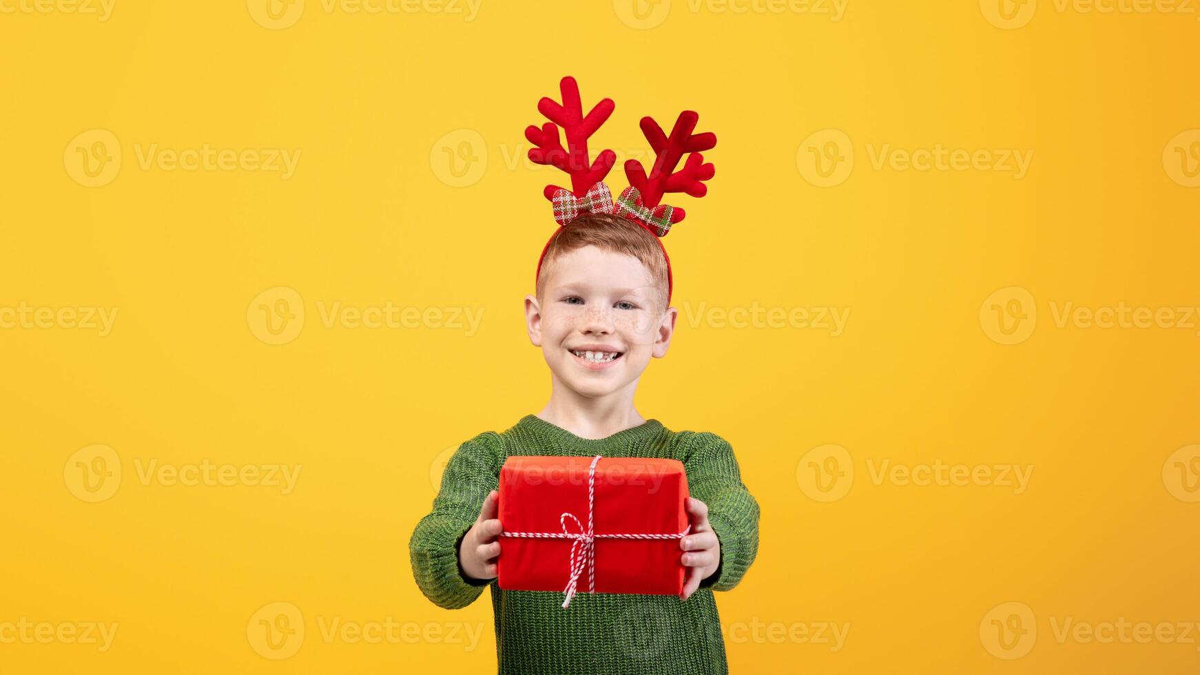 Smiling boy with festive deer accessory giving Christmas gift over yellow studio background, empty space. Happy kid celebrating New Year, cheerfully outstretching present to camera photo