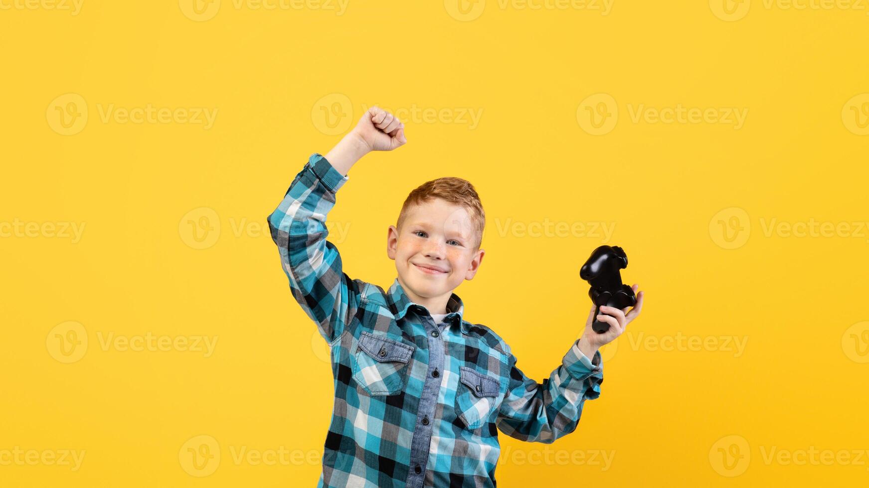 Happy boy playing game with joystick alone on yellow studio background, copy space. Emotional kid raising hands up, celebrating success, holding joystick, kids leisure concept photo