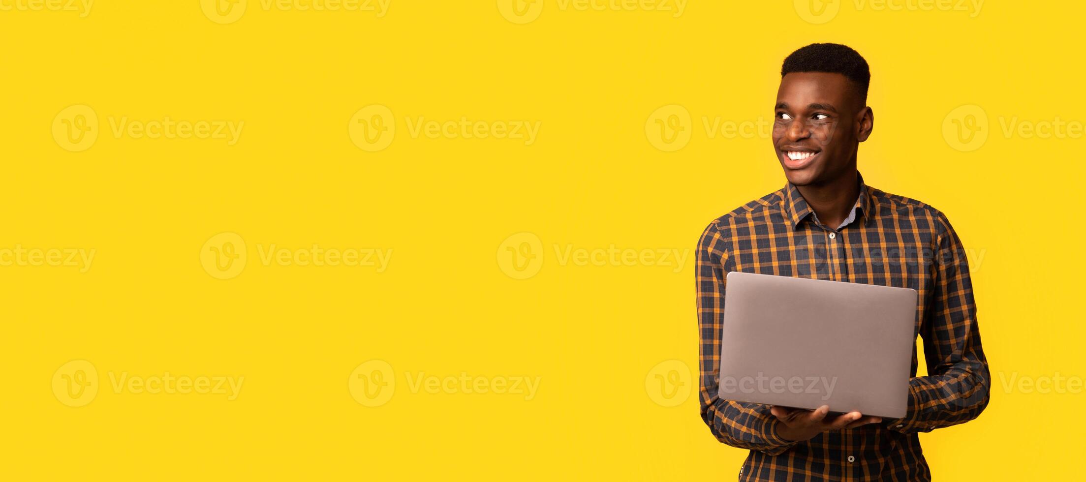 Remote job for students. Portrait of happy young black guy with laptop computer in hands looking aside at copy space, standing on yellow background, millennial African man enjoying freelance work photo