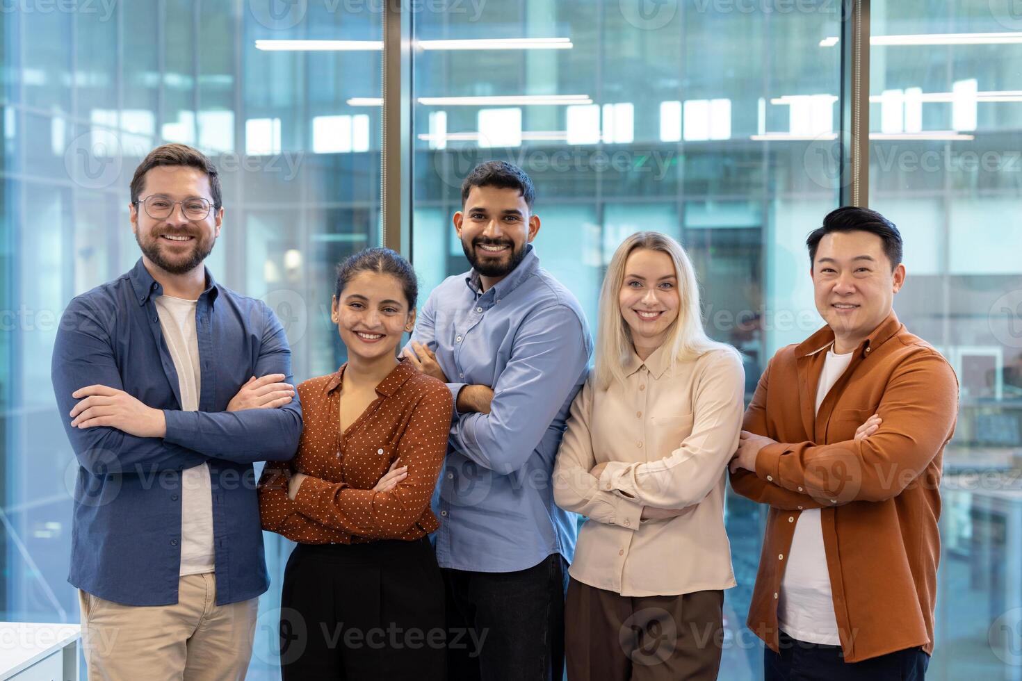 Diverse business team standing together, arms crossed, smiling confidently in an office setting with a modern glass wall. photo