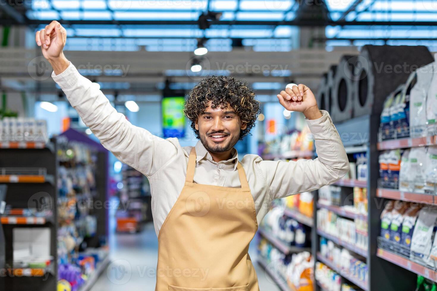 A cheerful store employee with curly hair celebrates with arms raised, smiling broadly in a supermarket aisle. photo