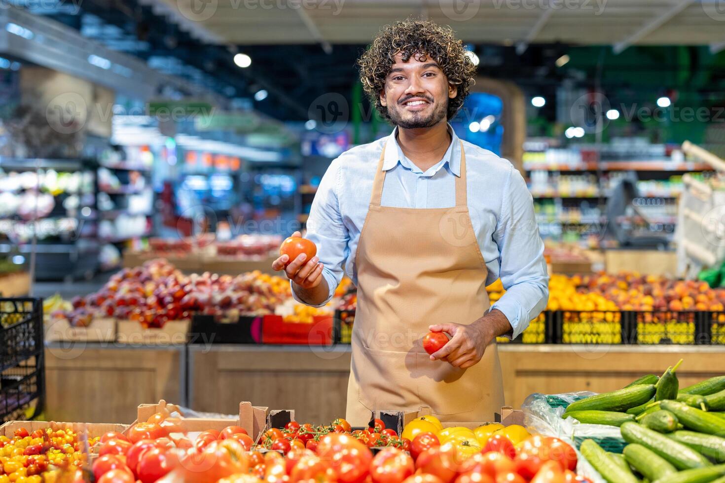 A smiling grocery store worker stands behind a counter, holding tomatoes and looking at the camera, with produce in the foreground. photo