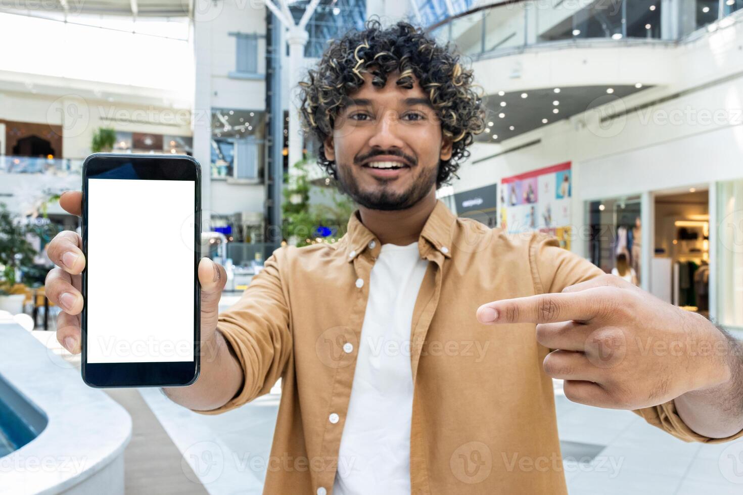 A smiling man with curly hair holds up a phone with a blank screen, pointing at it in a modern mall. photo