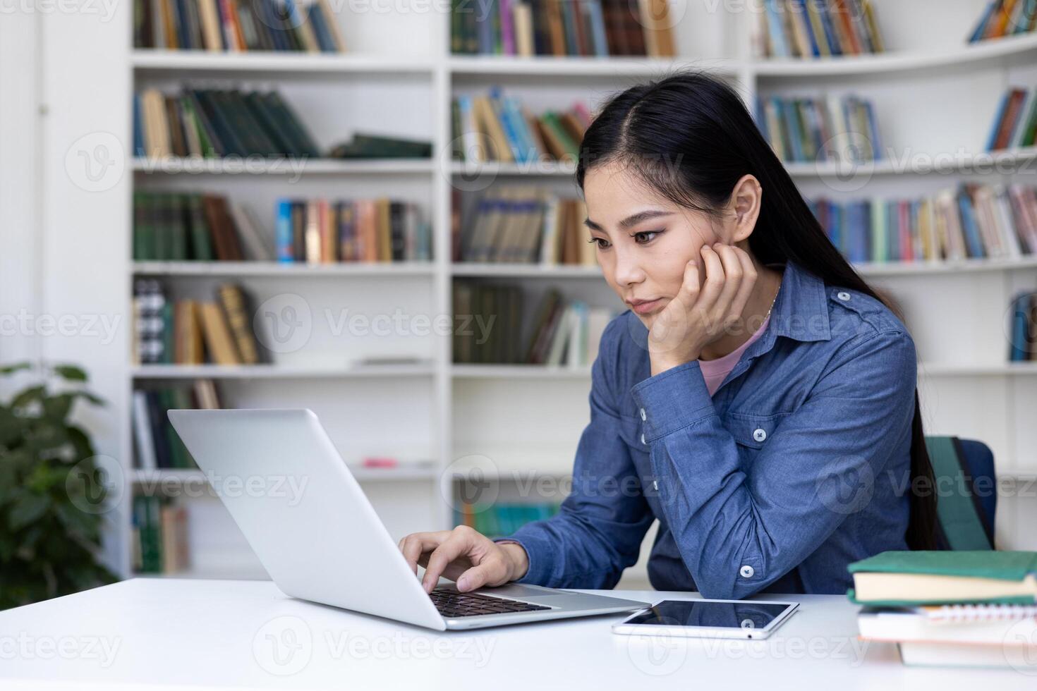 A focused Asian woman works on a laptop in a library, surrounded by books and a digital tablet. photo