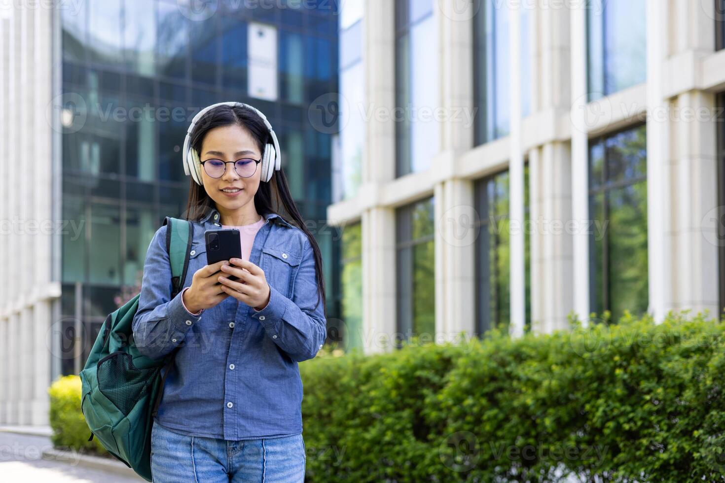 A young Asian woman in glasses and headphones looks at her phone while walking outside in an urban setting. photo