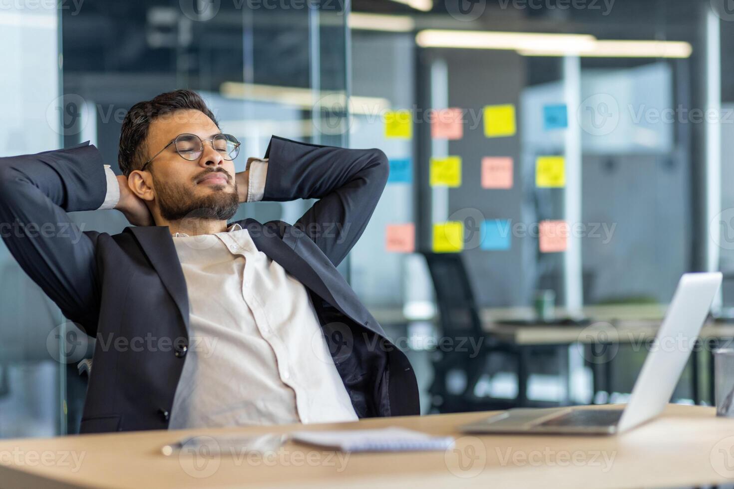 Businessman relaxing and resting inside the office. Man in business suit with hands behind head with closed eyes thinking and dreaming. Boss at workplace, working with laptop. photo