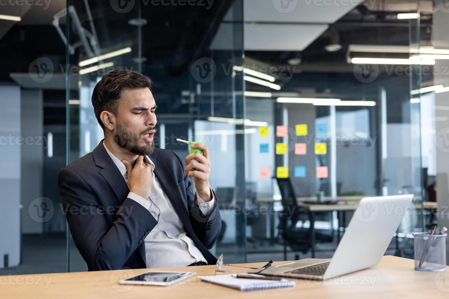 Man suffering from sore throat, company employee using spray inhaler medicine for relief. Businessman in business suit working inside office with laptop. photo