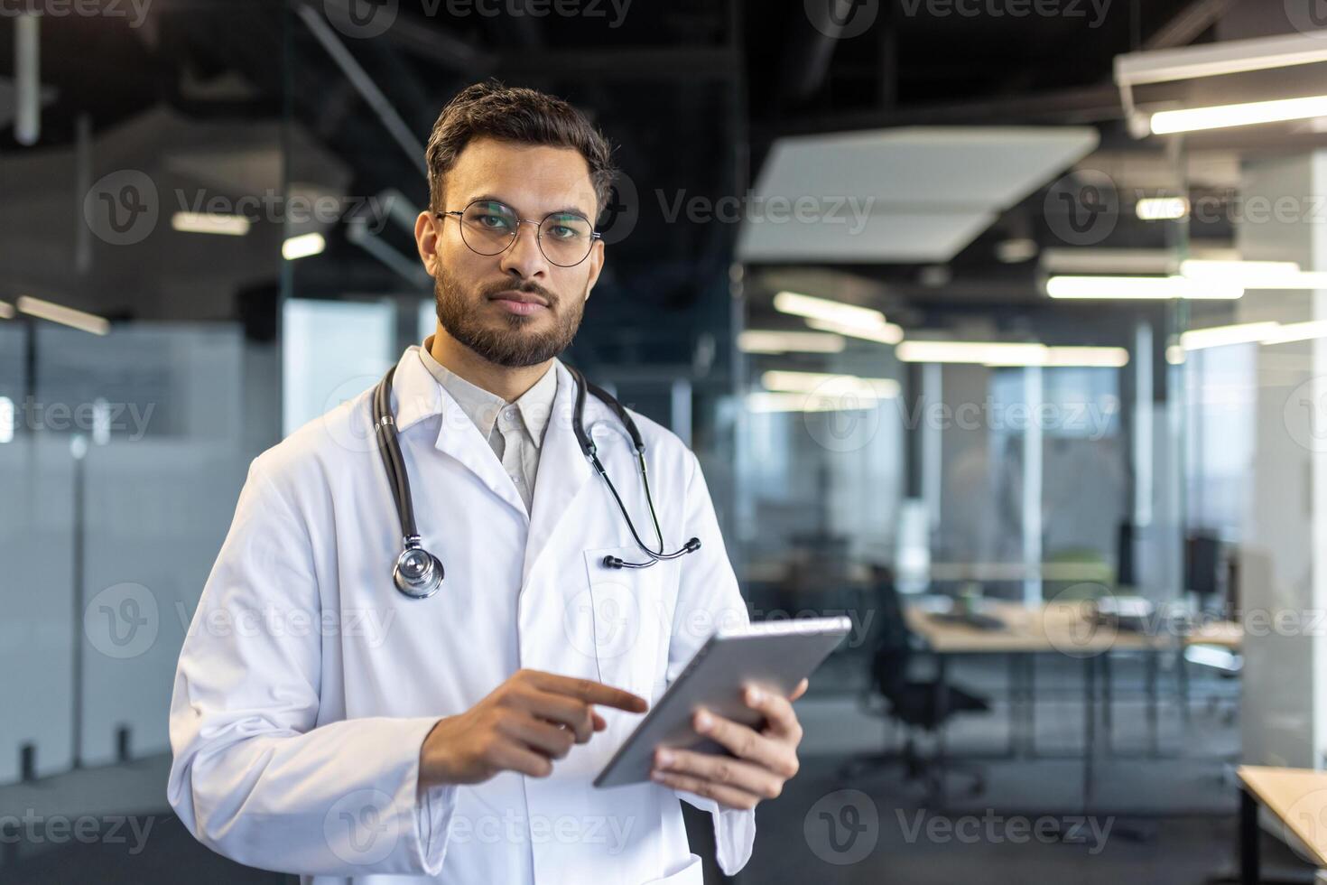 Portrait of serious thoughtful doctor, man in white medical coat and with stethoscope looking at camera. Medic uses tablet computer for online communication with patients. photo