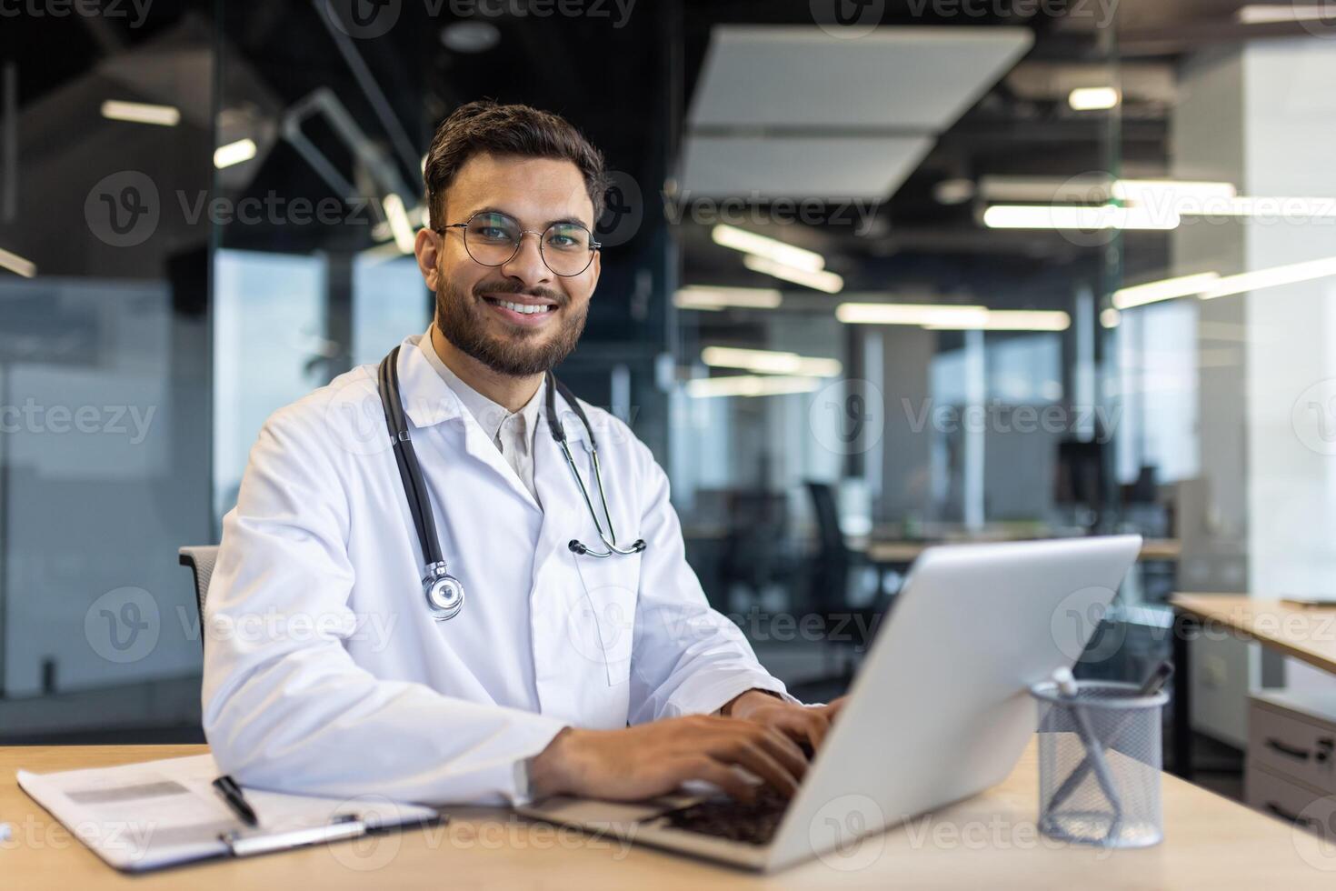 A smiling doctor, wearing a white coat and stethoscope, works on a laptop at a desk in a modern office setting. photo