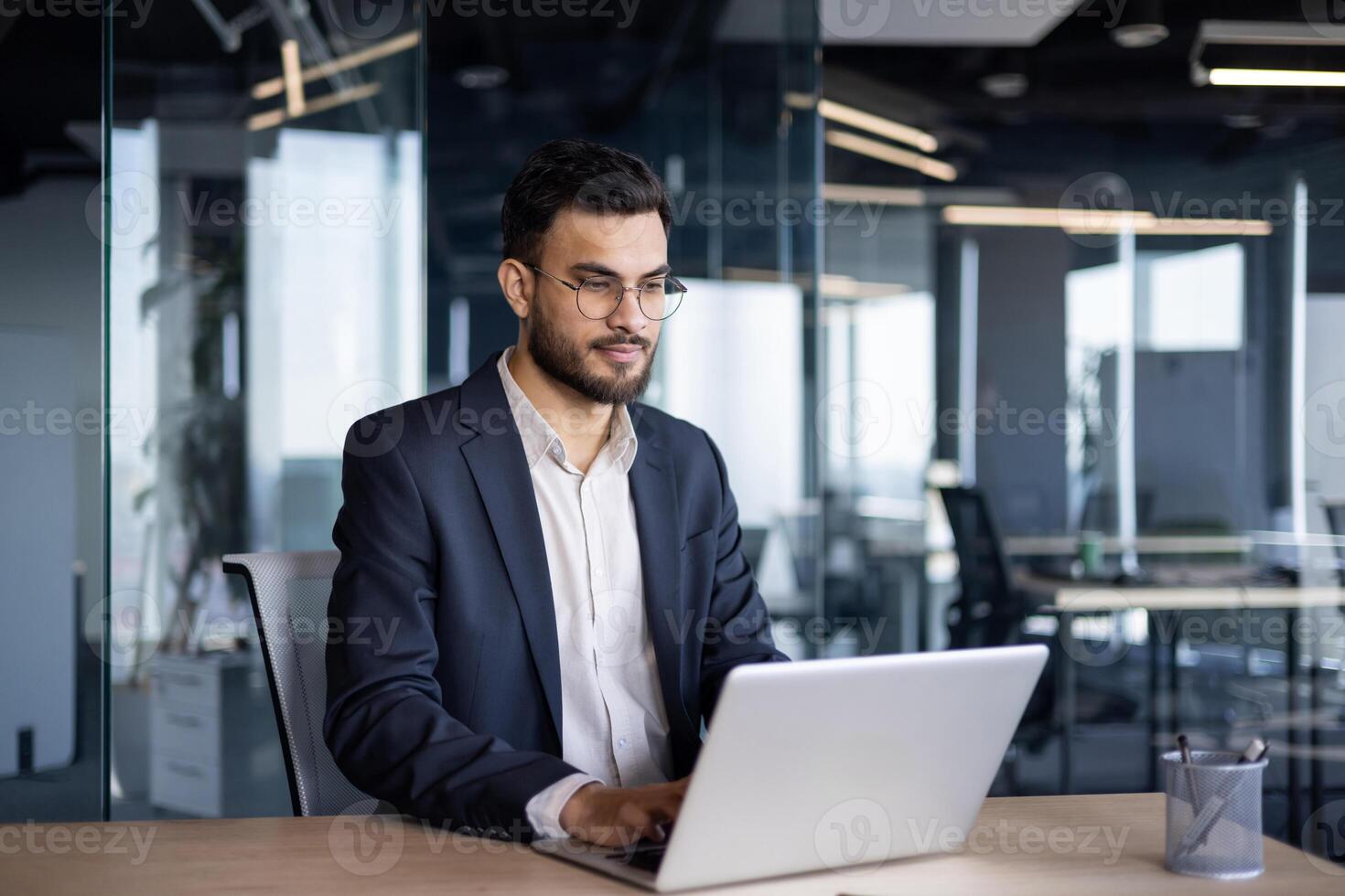 Serious thinking businessman working with laptop inside office. Man in business suit sitting at desk typing on keyboard, working late into the night. photo