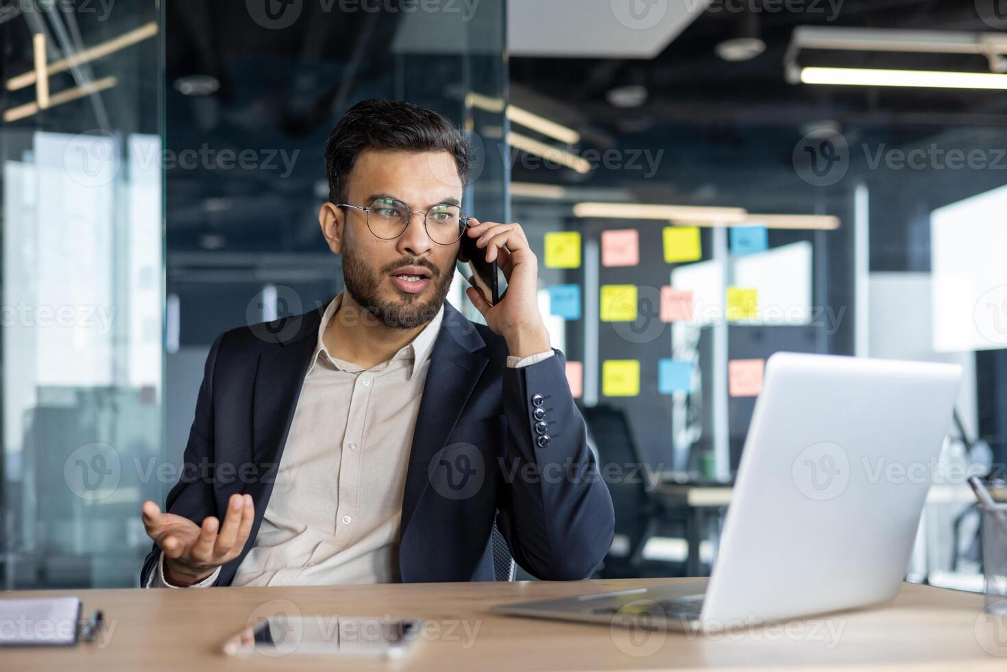 Serious and focused man talking on the phone inside the office. Businessman in business suit sitting at desk with laptop. photo