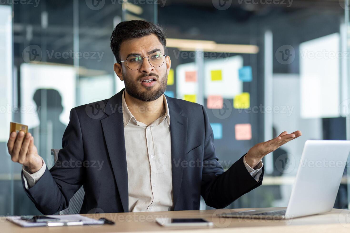 A confused businessman looks at a credit card in his hand while working on a laptop in an office environment. The man is expressing financial uncertainty. photo