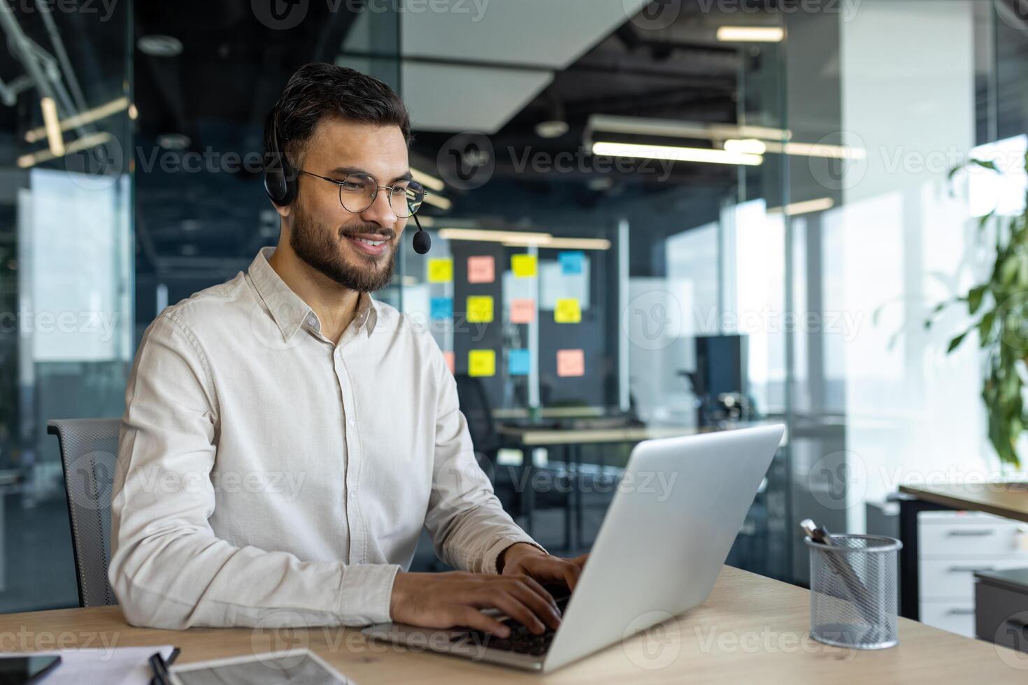 A smiling man with glasses and a headset works on a laptop at an office desk. He appears to be engaged in a call. photo
