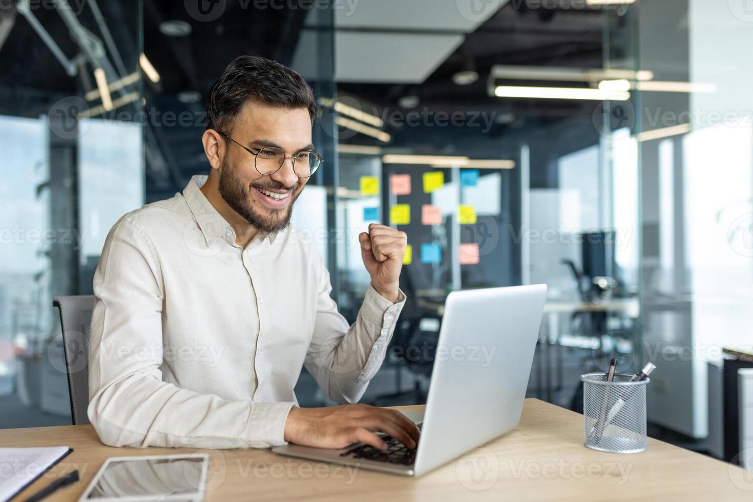 Man celebrating victory and successful achievement results at workplace. Businessman holding hand up happy smiling triumph gesture. Worker inside office working with laptop. photo