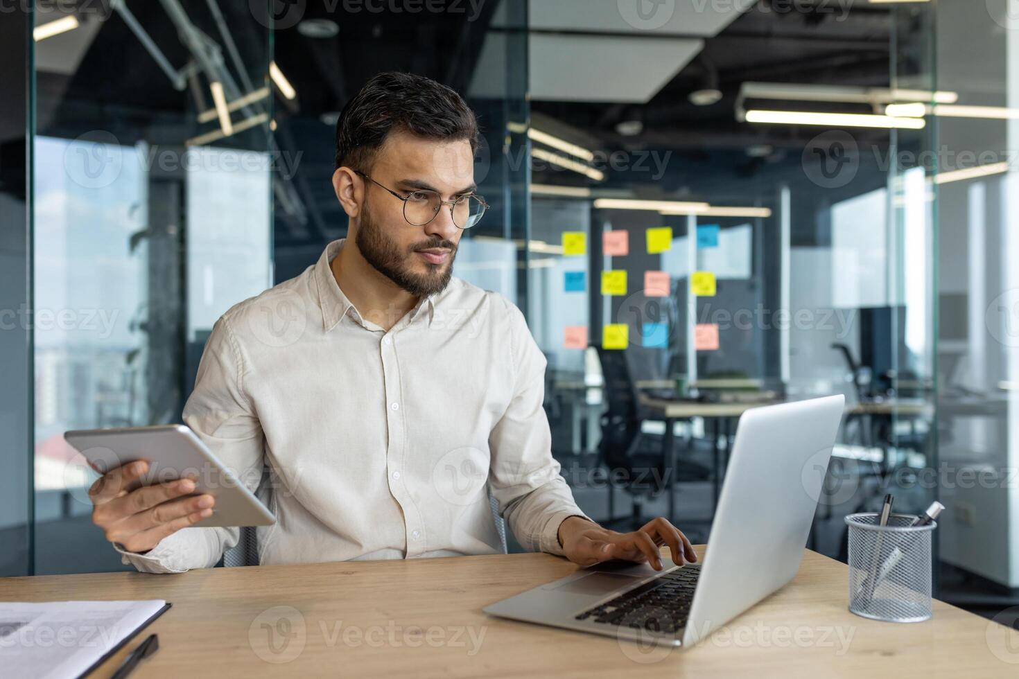 Multitasking, man with tablet computer working inside office using laptop. Businessman serious and focused typing on keyboard. Company employee thinking and concentrated. photo
