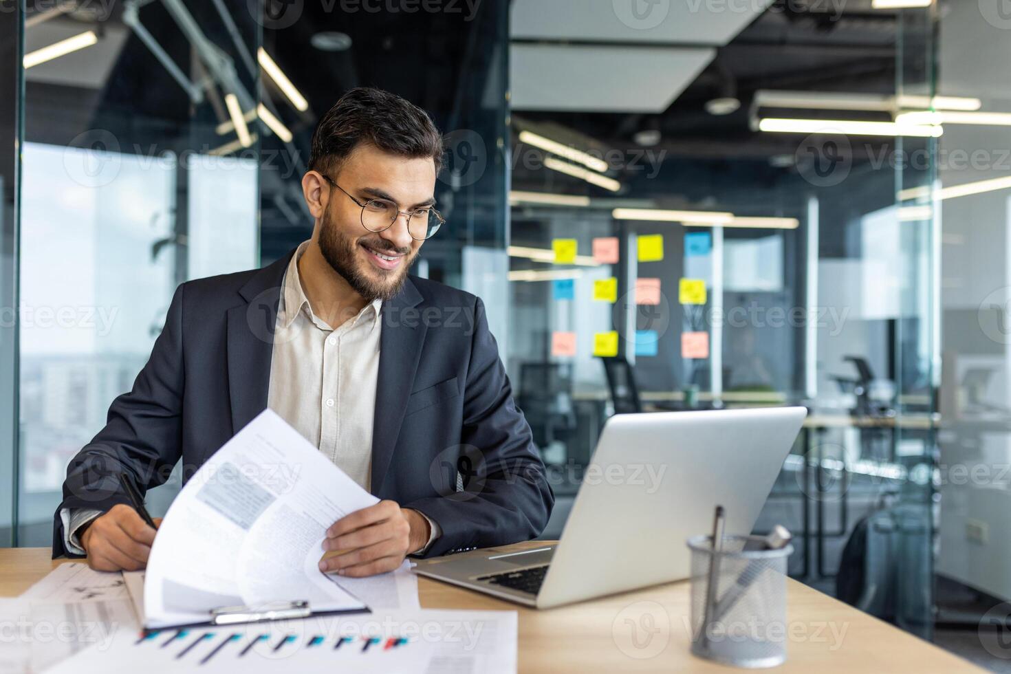 A smiling businessman reviews documents, with a laptop open on his desk in a modern office setting, ready to work. photo