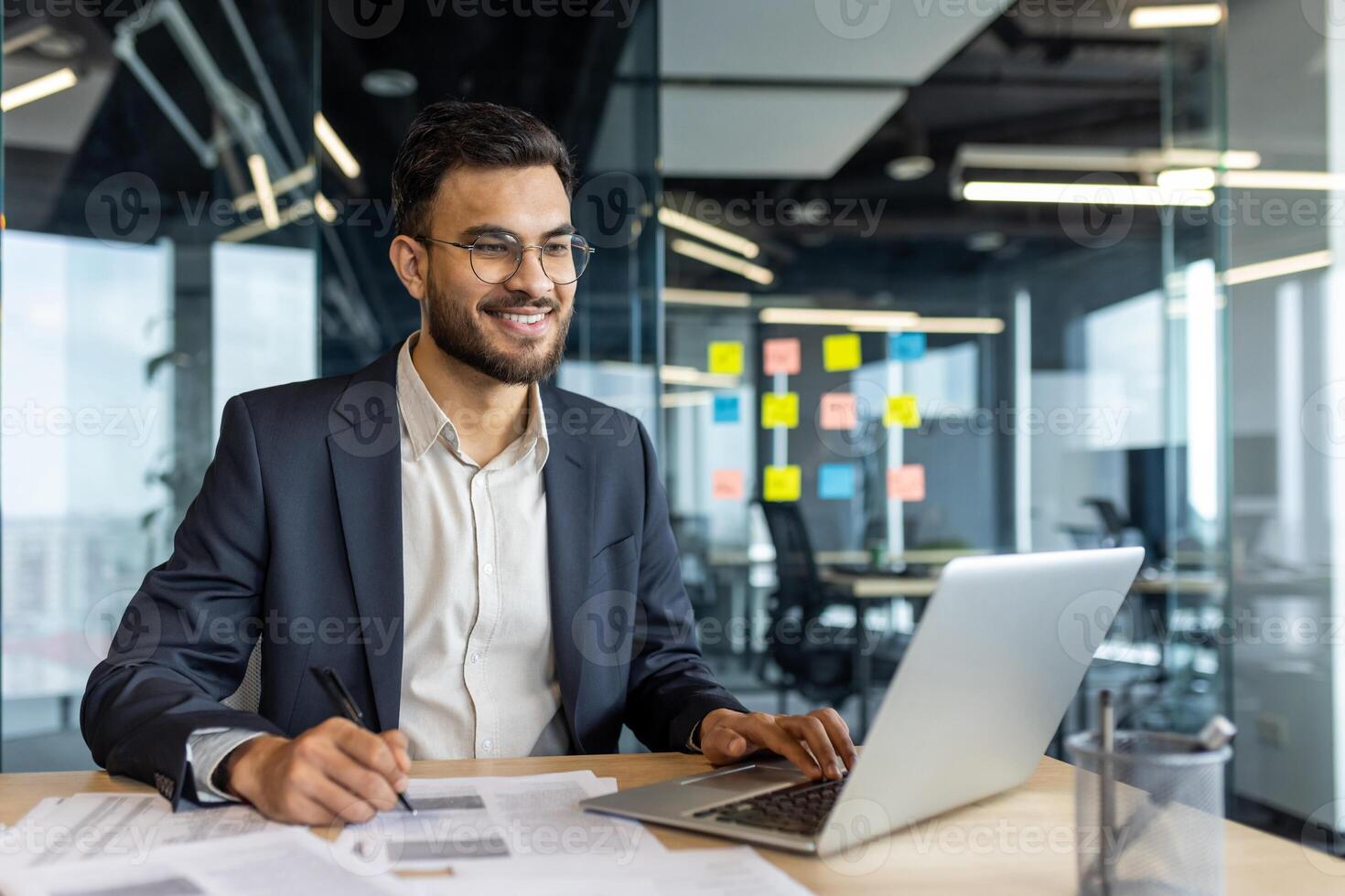 A smiling businessman in glasses works on a laptop and documents in a modern office setting, suggesting productivity and business success. photo